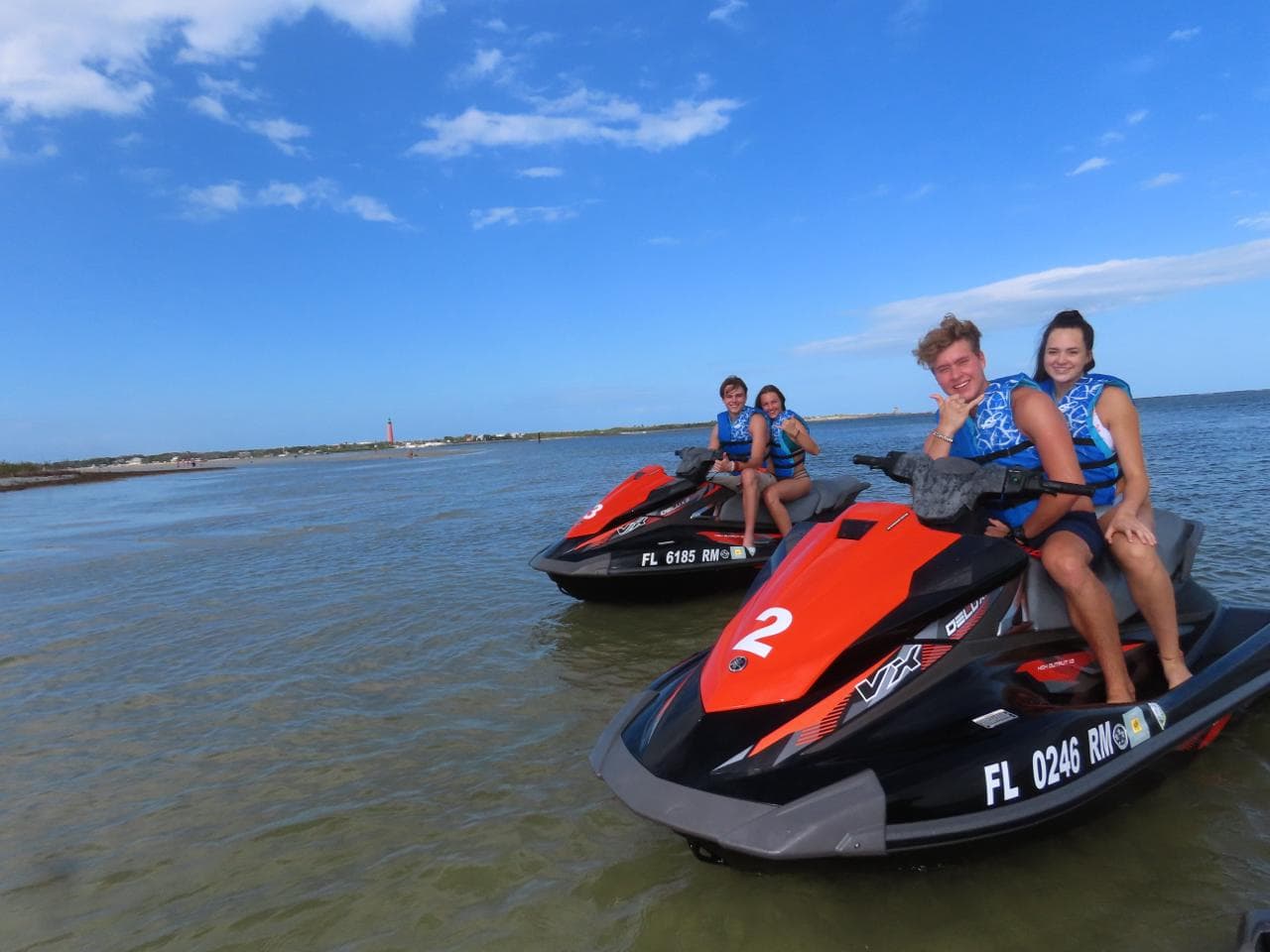 Four guests on two jet skis grinning at the camera in the shallows, the lighthouse small on the horizon.