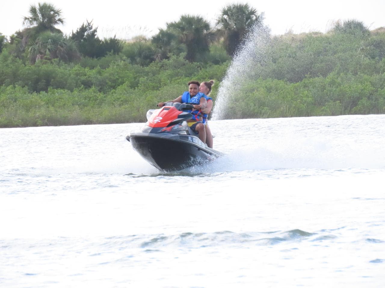 Two riders laughing as their jet ski throws spray in the estuary, mangroves along the bank.