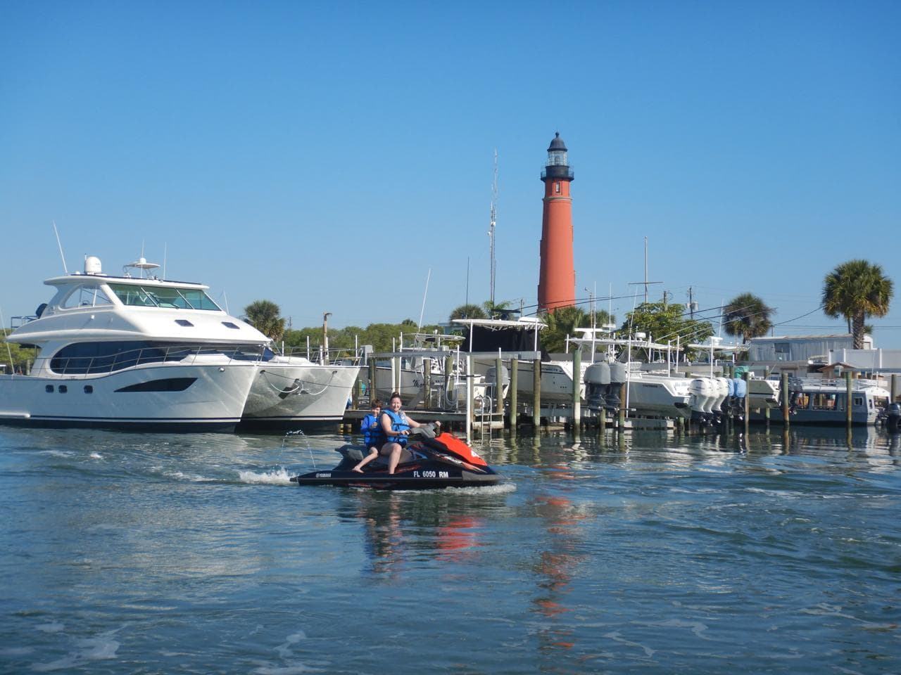 Two riders on a Yamaha WaveRunner idling past the marina with the red Ponce Inlet Lighthouse rising behind them.