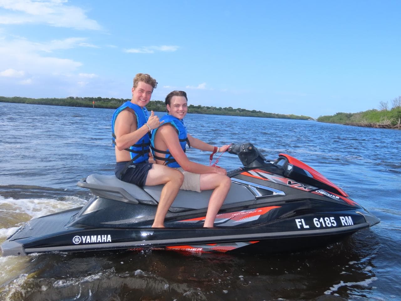 Two riders sitting on a jet ski in the estuary, giving a shaka to the camera.