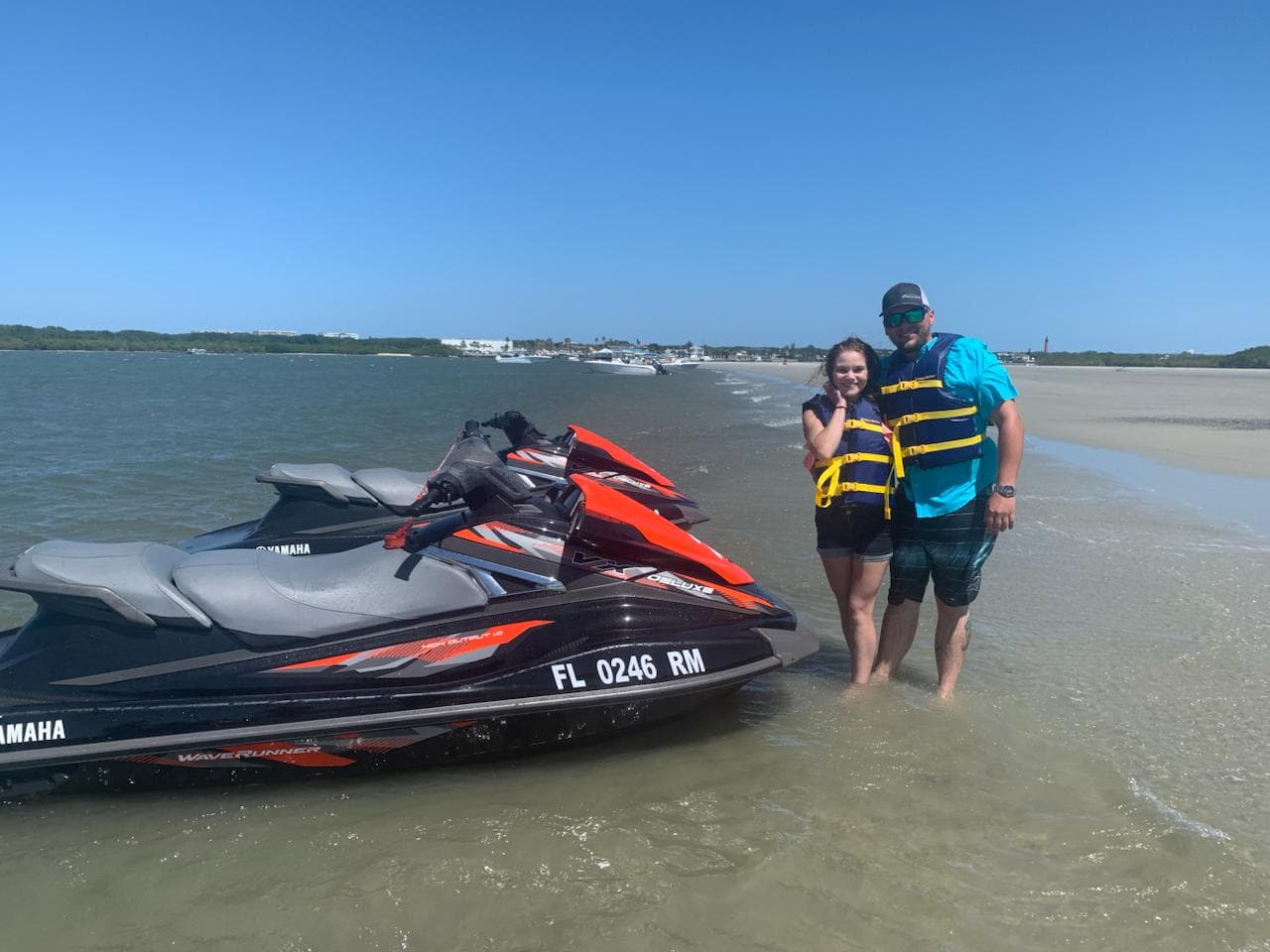Two guests standing in ankle-deep water beside their jet skis on a sandbar, life vests on.